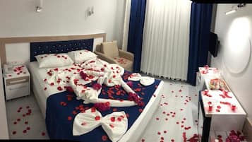 A hotel room with a bed covered in rose petals and a towel with a bow, a bedside table with a bottle, and a chair.