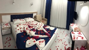 Una habitación de hotel con una cama cubierta de pétalos de rosa y una toalla adornada con un lazo, una mesita de noche con una botella y una silla.