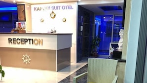 A reception area with a sign for Kapitan Suit Hotel, a reception desk, a ship's wheel table, and seating with yellow cushions. - Kaptan Suit Otel (Silifke)