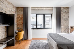 A modern hotel room with exposed brick walls, a large window with curtains, a bed, a desk with a chair, and a flat-screen TV mounted on the wall. - Porta 20 Boutique Guesthouse (Leiria)