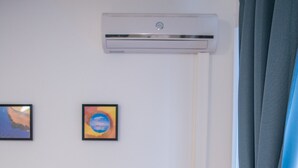 Une pièce de taille modeste, dotée d’un bureau en bois, d’un meuble de rangement blanc, d’un climatiseur et d’une fenêtre avec des rideaux.