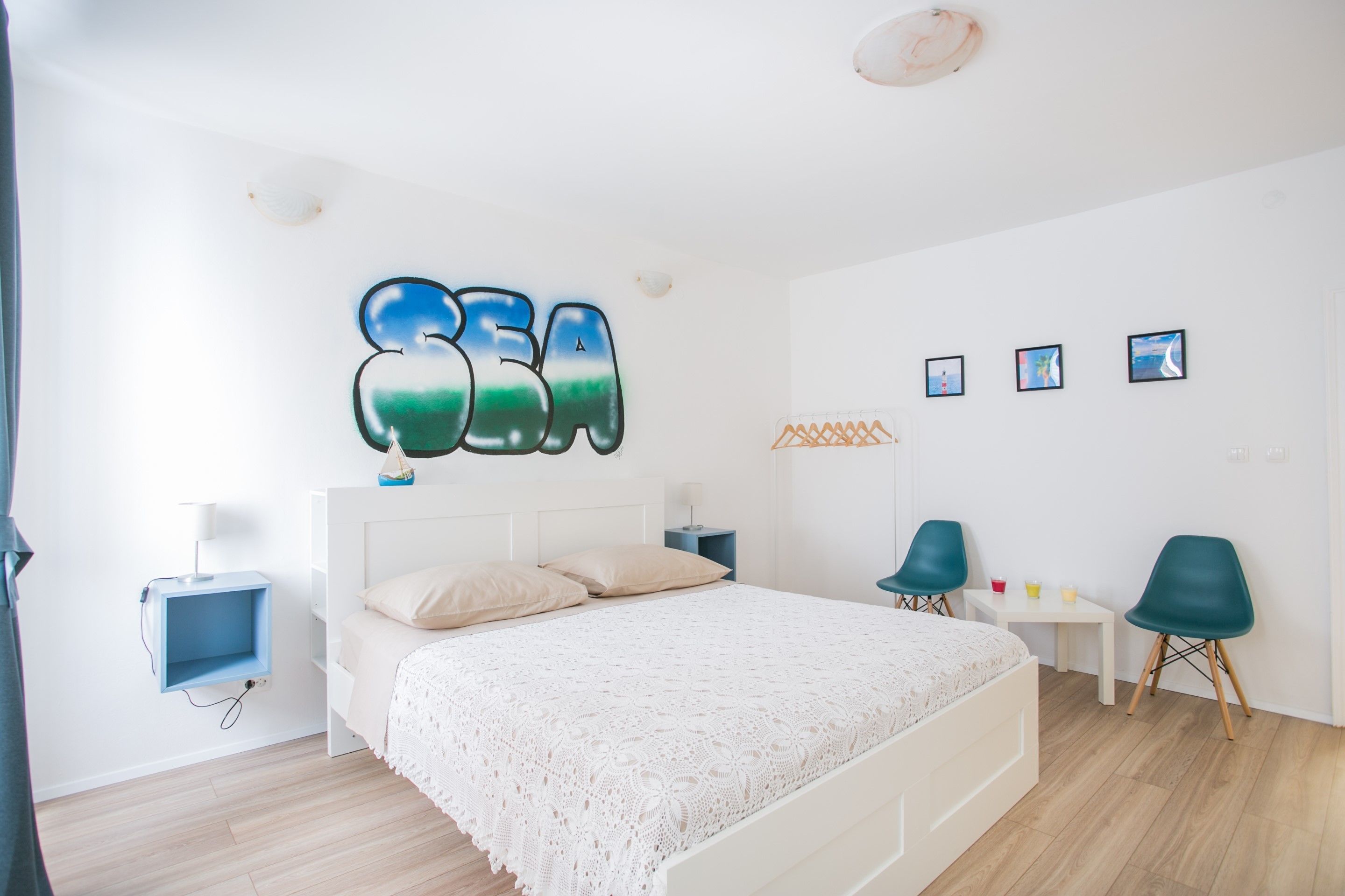 Un dormitorio con cama, escritorio, dos sillas, una estantería montada en la pared y un adorno mural que dice 'SEA'.