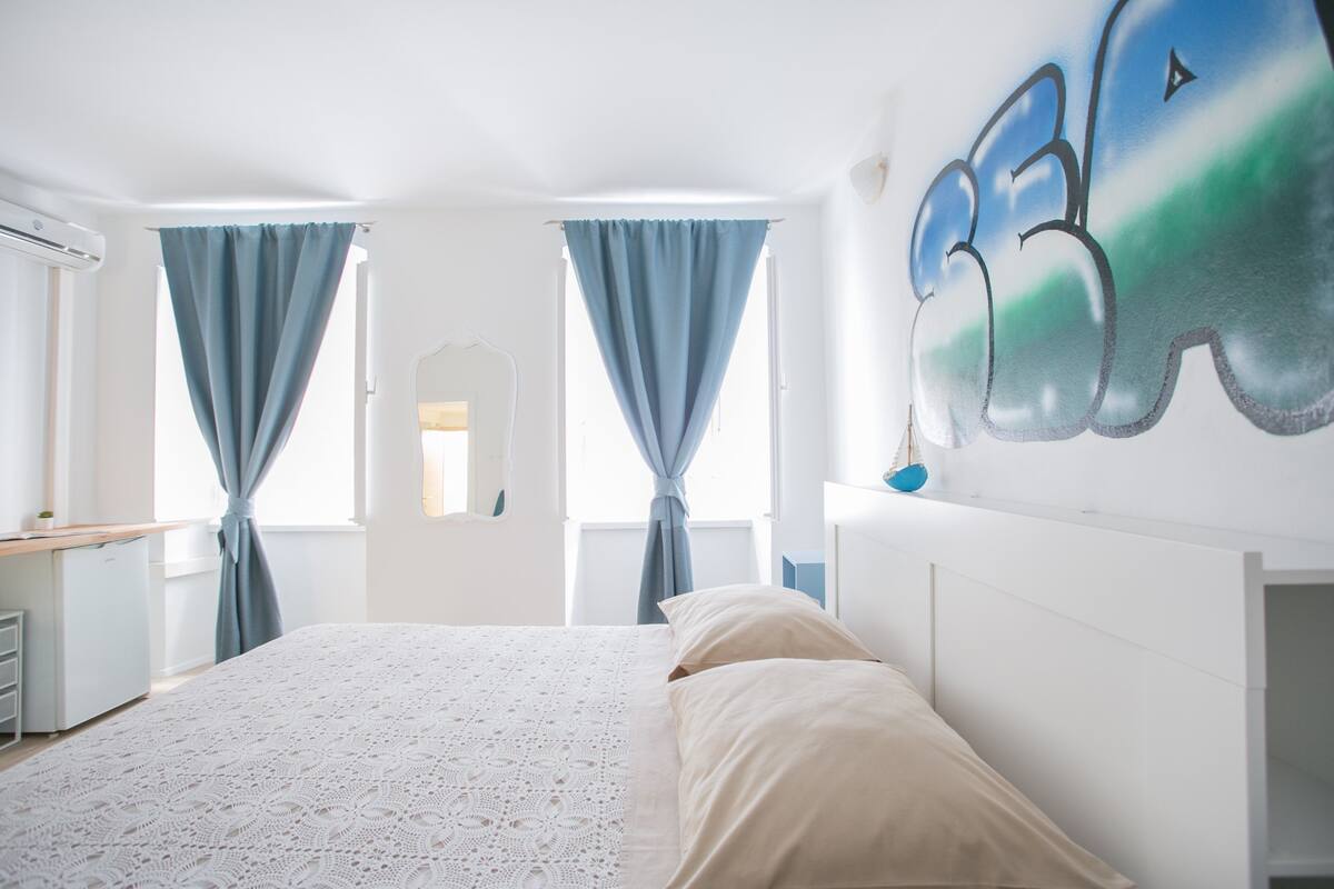 Un dormitorio con cama, cortinas, una pared con un cuadro y un escritorio con silla.