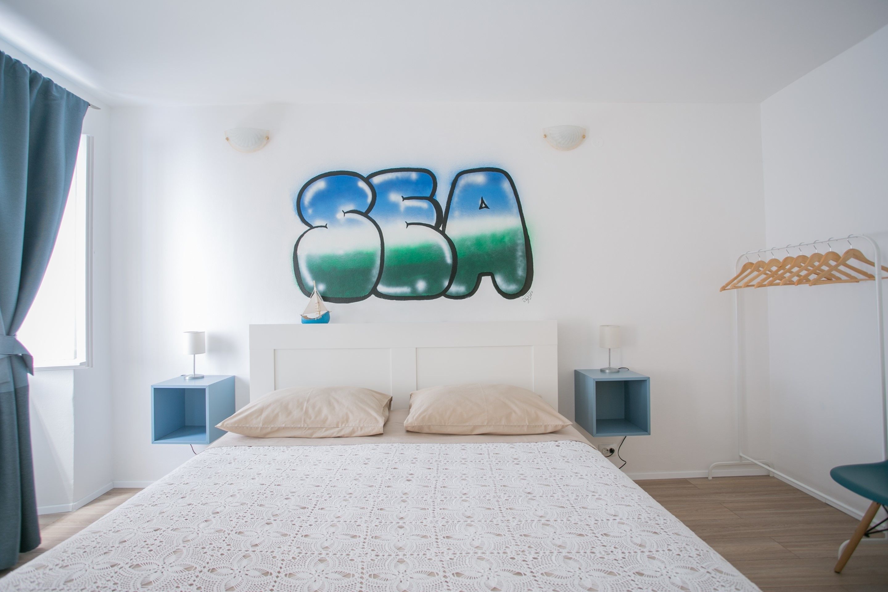 A bedroom with a bed, two bedside tables, a wall-mounted artwork, a wardrobe, and a hanging clothes rack.