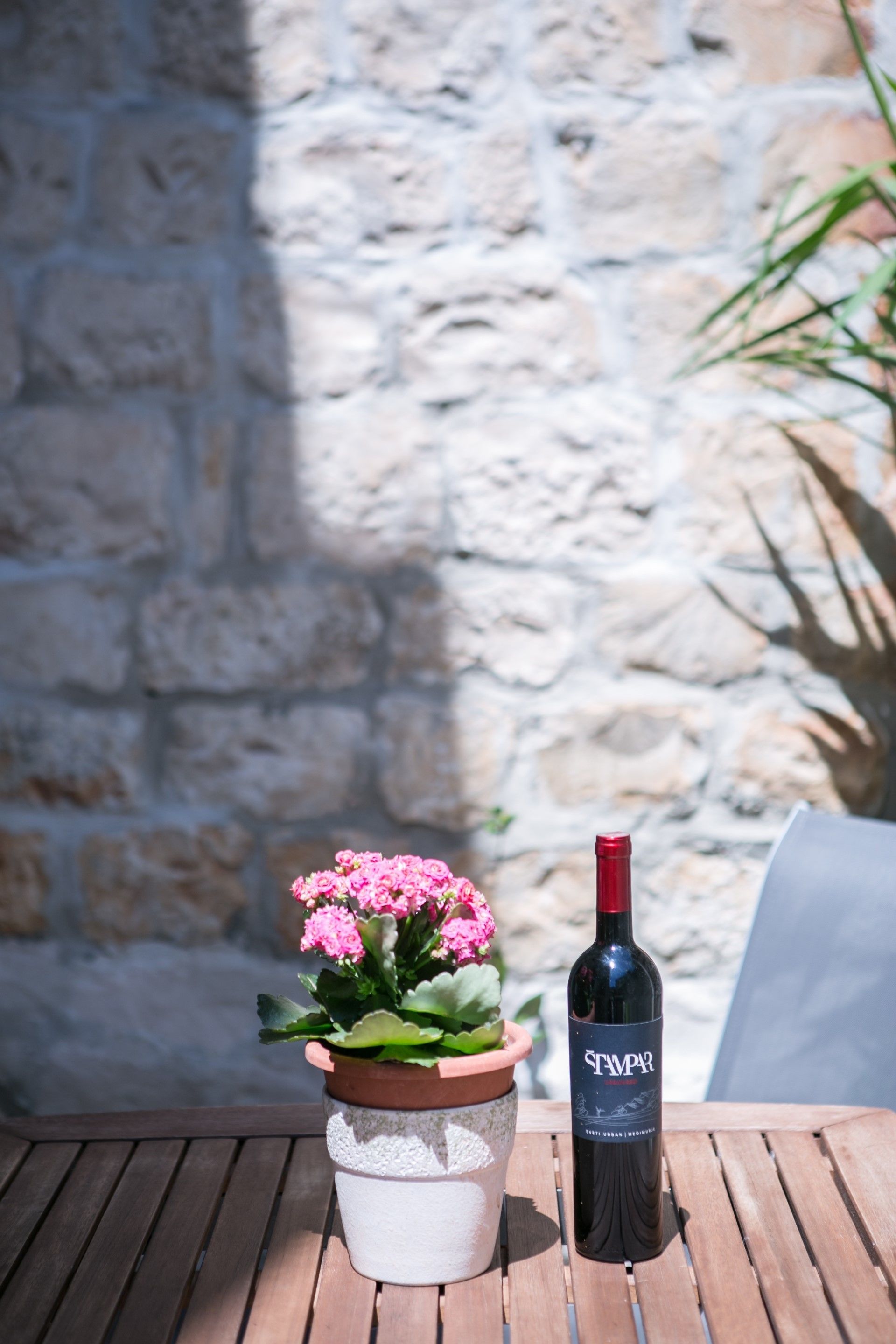 A bottle of wine and a potted plant on a wooden table against a stone wall.