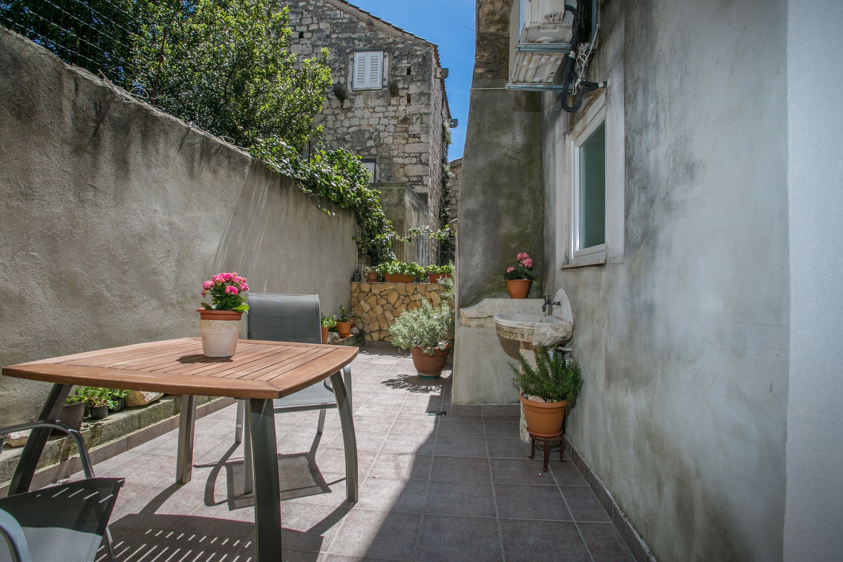 A narrow outdoor patio with a wooden table and chairs, surrounded by potted plants and a stone wall.
