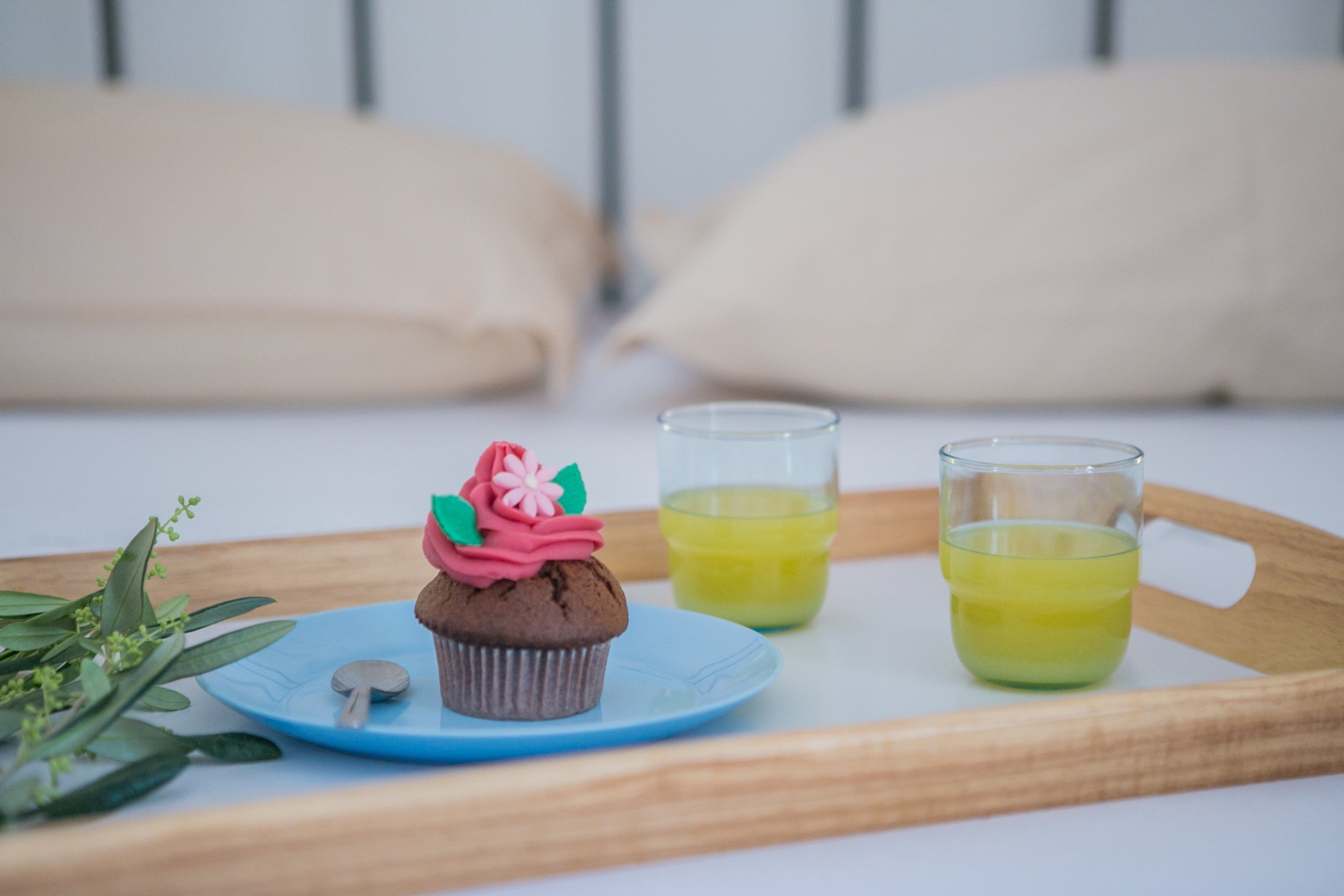 Ein Tablett mit einem Cupcake und zwei Gläsern gelber Flüssigkeit auf einem Bett mit beige farbenen Kissen.