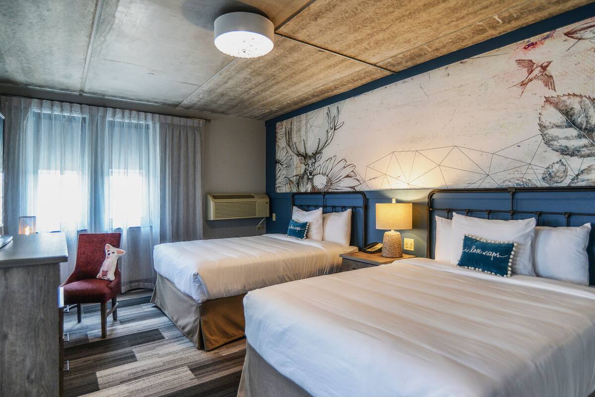 Habitación de hotel con dos camas, un escritorio, una silla y un mural grande en la pared.