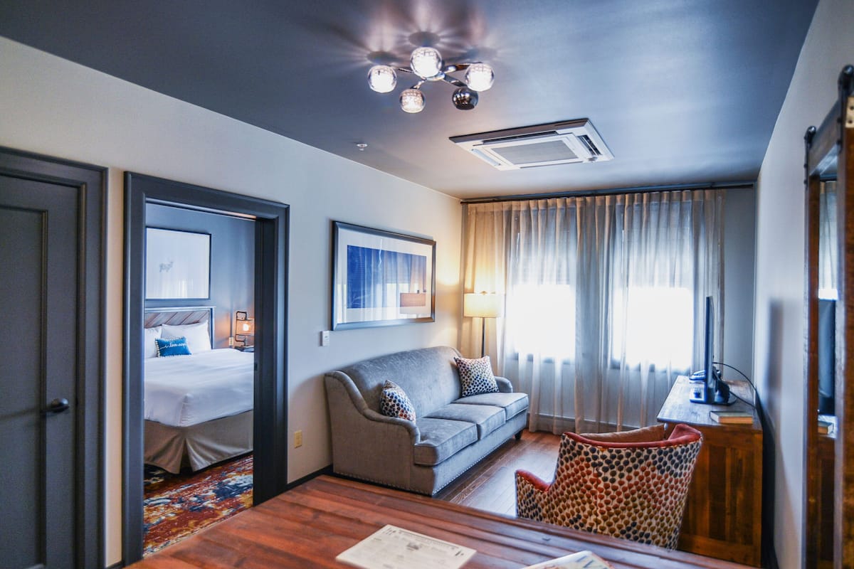 Ein Hotelzimmer mit einem Bett, einem grauen Sofa, einem Holztisch mit Zeitschriften, einem gemusterten Sessel, einem Schreibtisch mit einem Computer und einem Fenster mit Vorhängen.