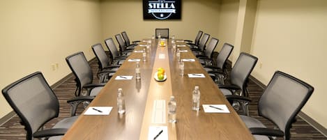 Un salón de conferencias con una larga mesa, sillas, botellas de agua y un plato de fruta.