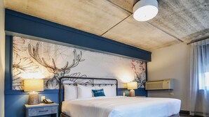 A hotel room with a large bed, two bedside tables with lamps, a wall-mounted air conditioning unit, and a decorative wall piece. - The Stella Hotel & Ballroom (Kenosha)