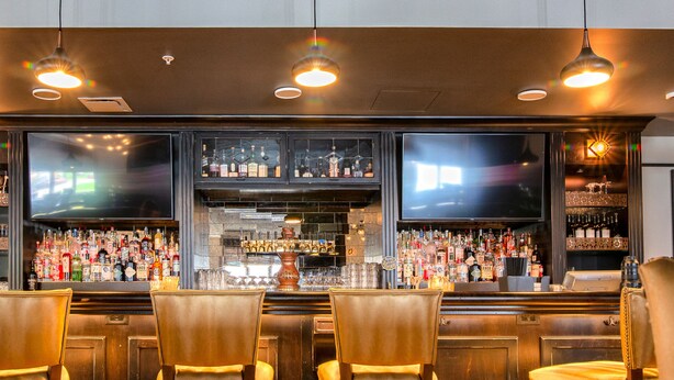 A bar area with multiple seating options, a television screen, and a variety of bottles and glasses on shelves. - The Stella Hotel & Ballroom (Kenosha)