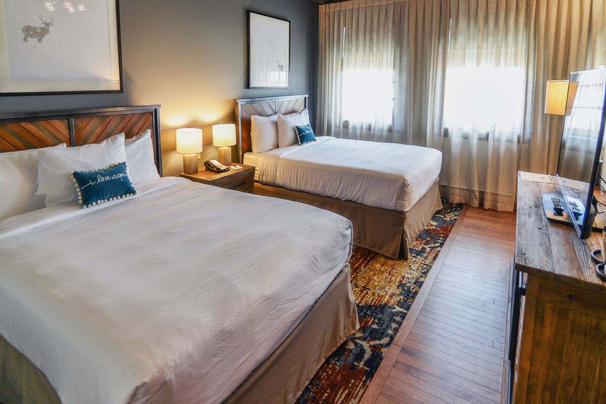 Ein Hotelzimmer mit zwei Betten, einem hölzernen Kopfteil, einem Nachttisch mit Lampe, einem Schreibtisch mit Computer und einem großen Fenster mit Vorhängen.