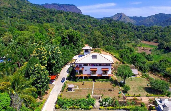Uma casa de dois andares com telhado plano e uma pequena construção anexa, rodeada por vegetação exuberante e colinas ao fundo.