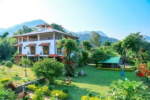 Um edifício de dois andares com um jardim na frente, rodeado por vegetação exuberante e montanhas ao fundo.