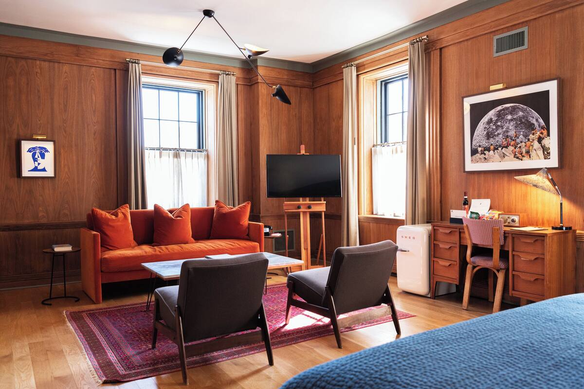 A hotel room with a wooden paneled wall, a large flat-screen TV, an orange sofa, two chairs, a desk, and a bed.