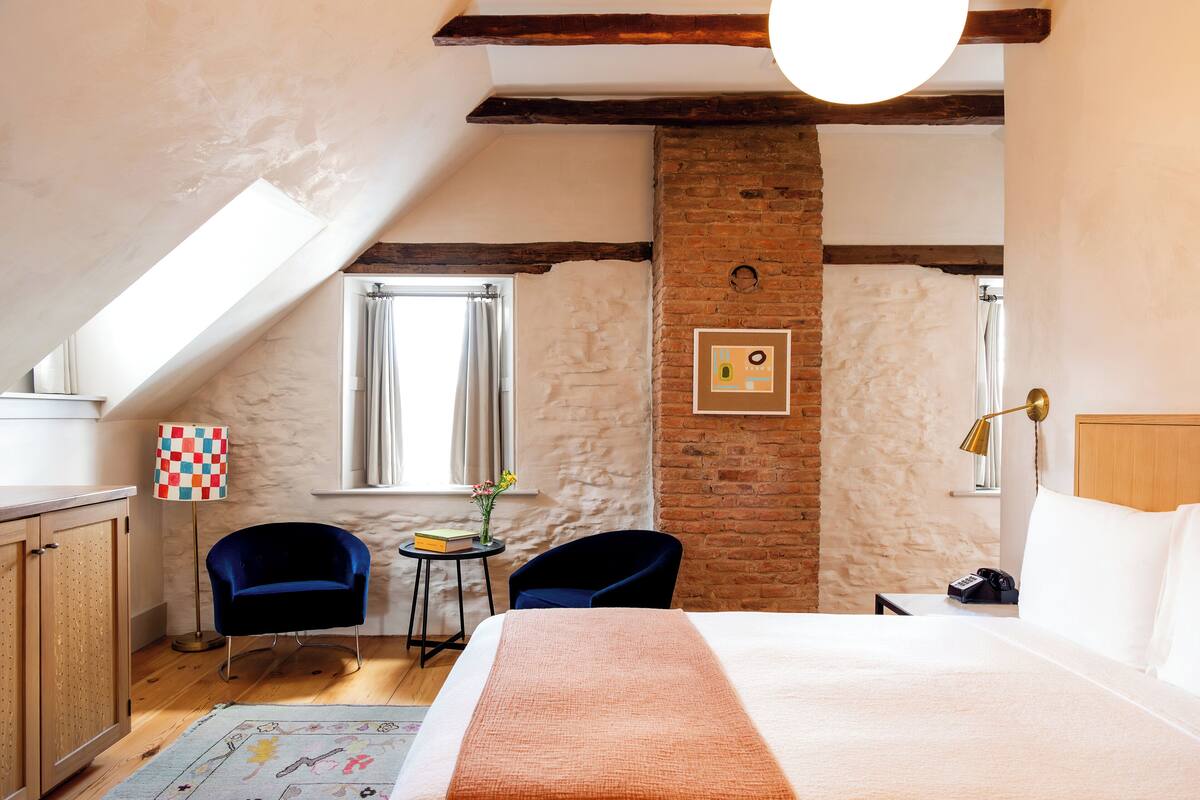A hotel room with a sloped ceiling, exposed brick walls, a large bed, two chairs, a small table with a lamp, and a window with curtains.