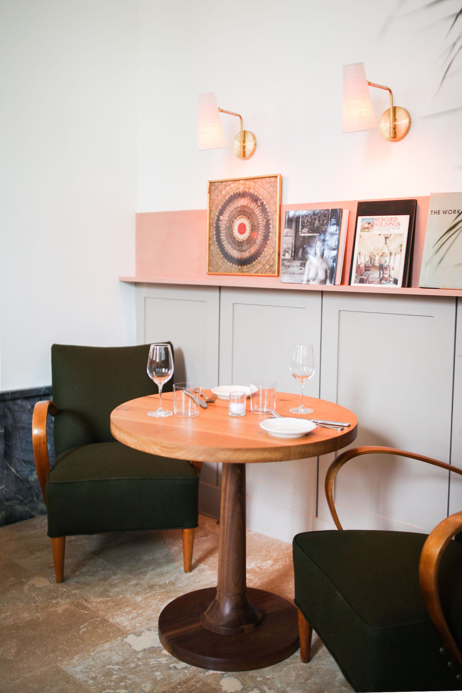 A dining area with a round wooden table, two green upholstered chairs, and a small round table with a single chair. There are two wall-mounted lamps, a framed picture, and a book titled "The Work" on the wall.