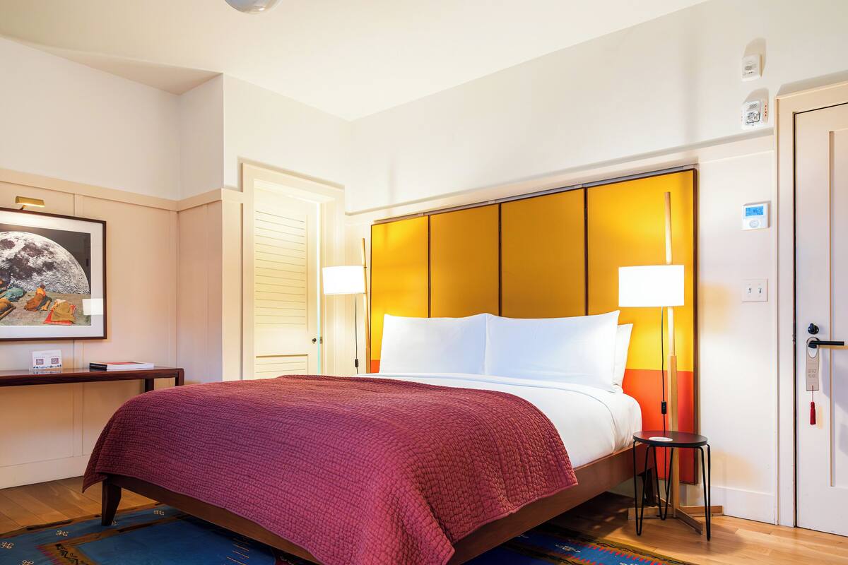 A hotel room with a bed, a nightstand, a lamp, a door, and a wall-mounted thermostat.