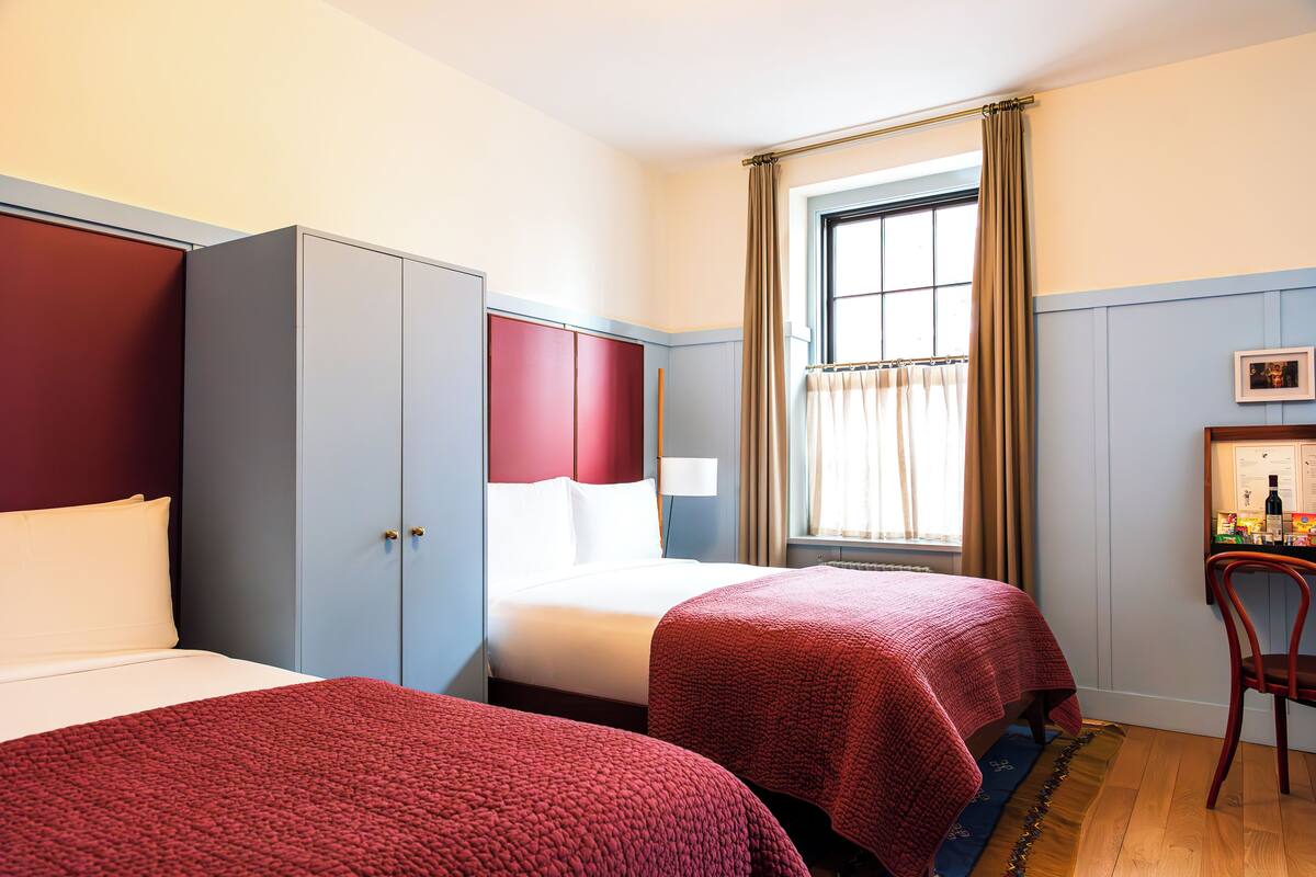 A hotel room with two beds, a wardrobe, a desk with a chair, and a window with curtains.