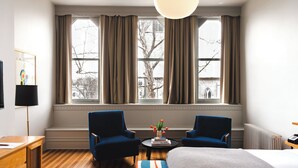 Een hotelkamer met een groot raam, twee blauwe fauteuils, een ronde tafel met een plant en een bed met een witte deken.
