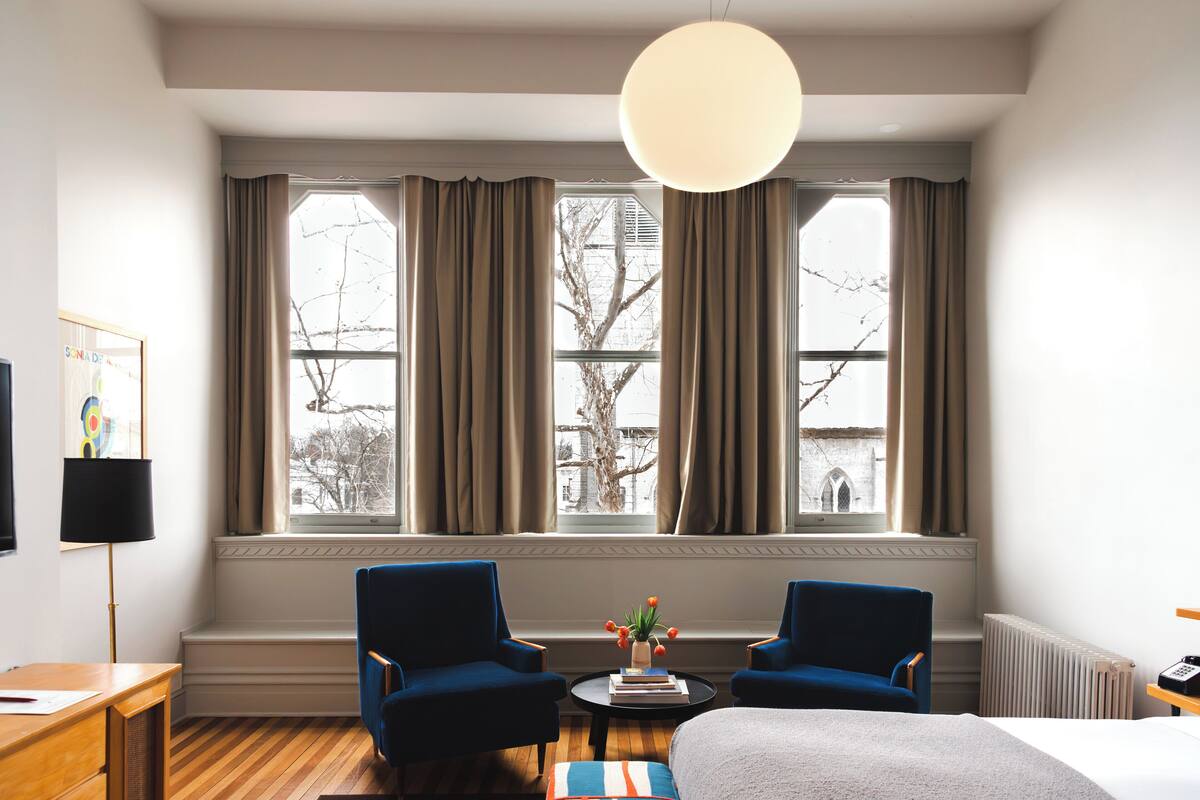 A hotel room with a large window, two blue armchairs, a round table with a plant, and a bed with a white blanket.
