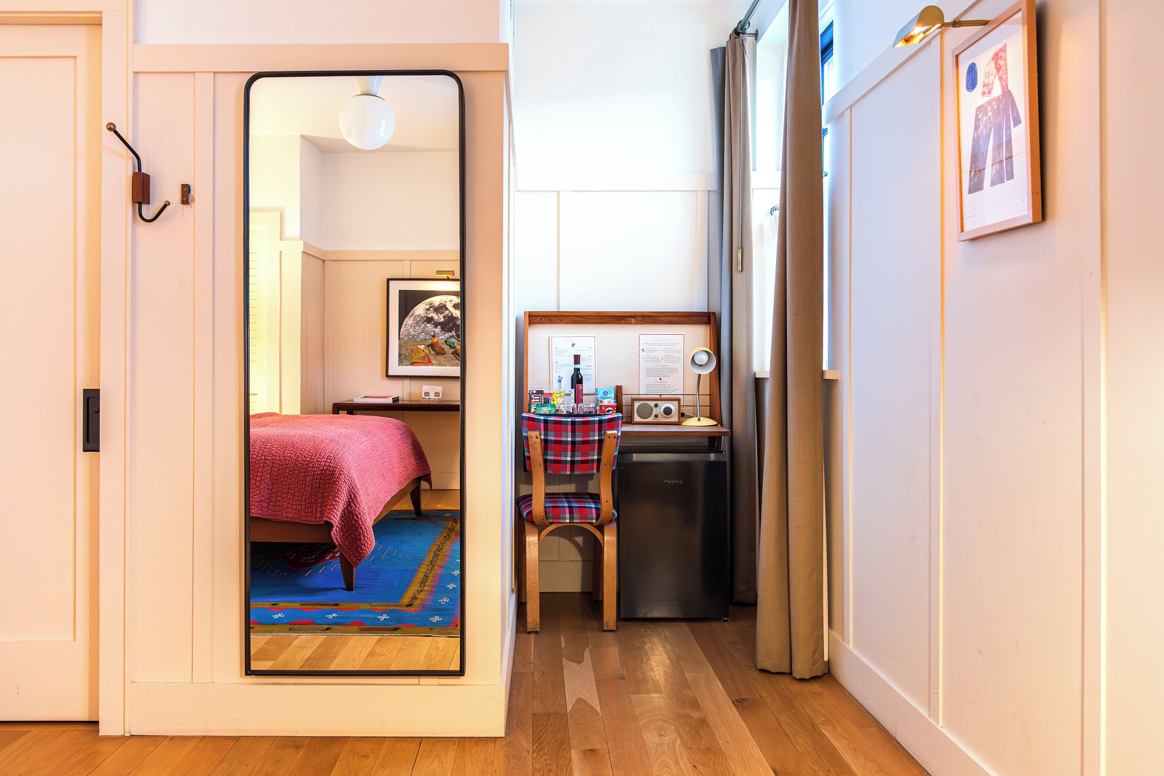 A compact room with a bed, a small table with a chair, a refrigerator, and a mirror reflecting another room with a bed and a rug.