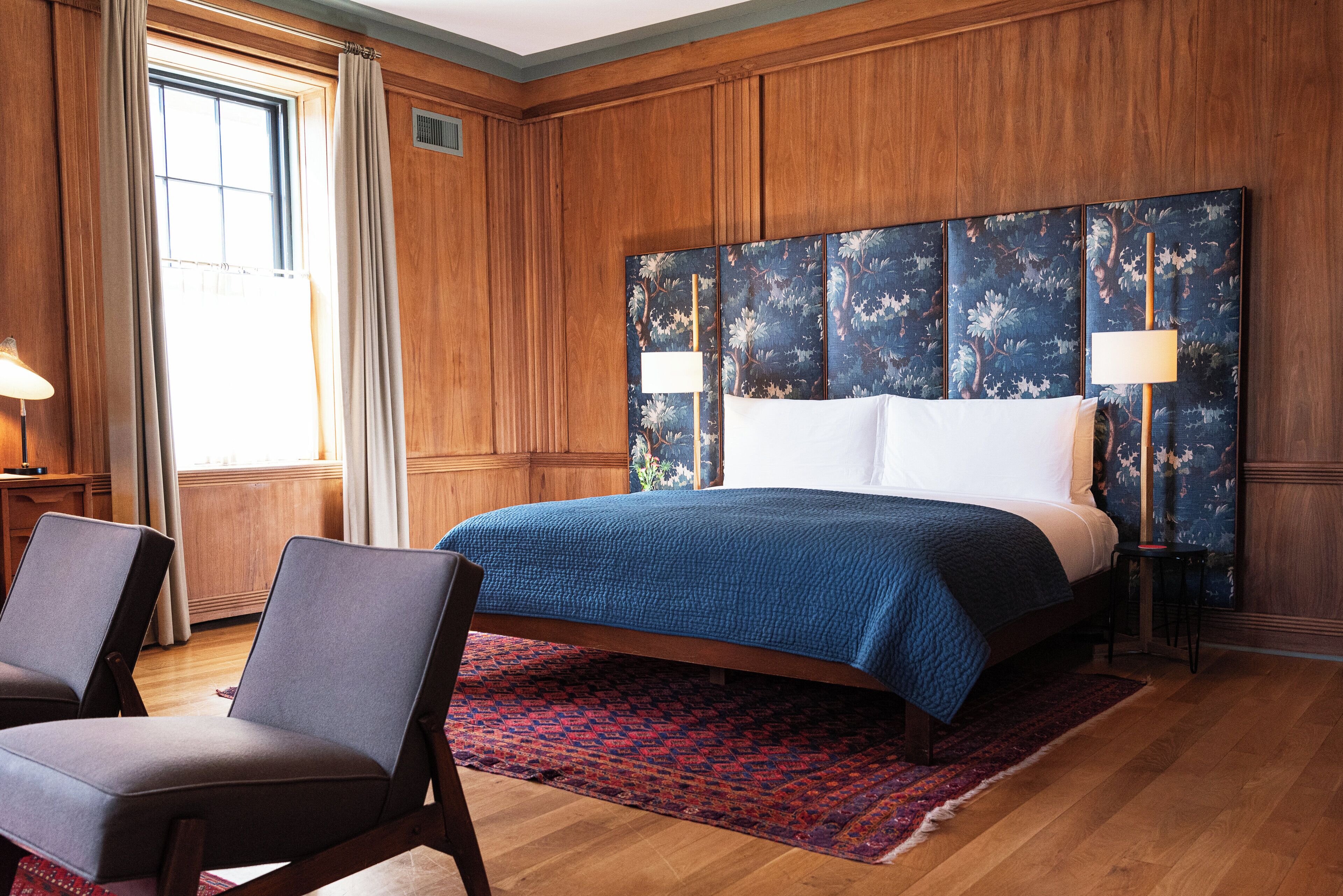 Une chambre d’hôtel avec un grand lit, deux chaises, une tête de lit en bois et un tapis rouge à motifs.