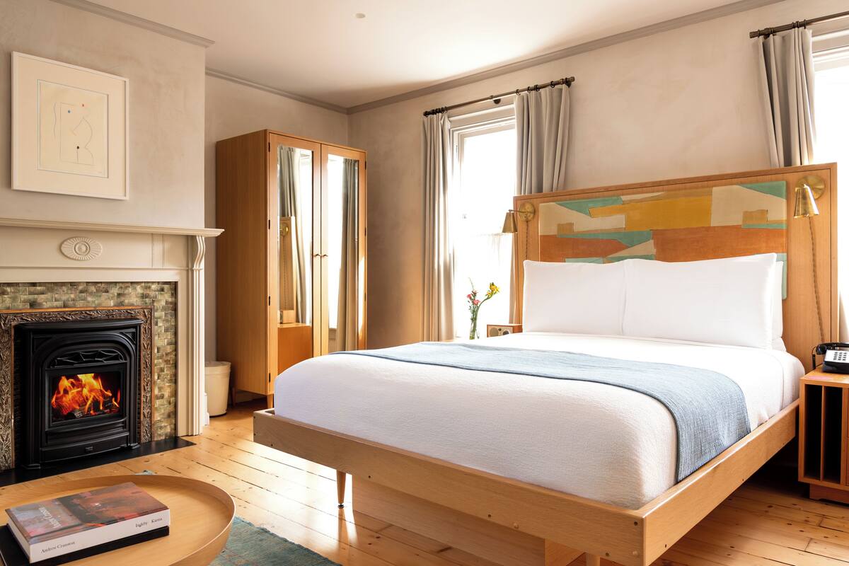 A bedroom with a wooden bed, a fireplace, a wooden wardrobe, and a bedside table with a book.