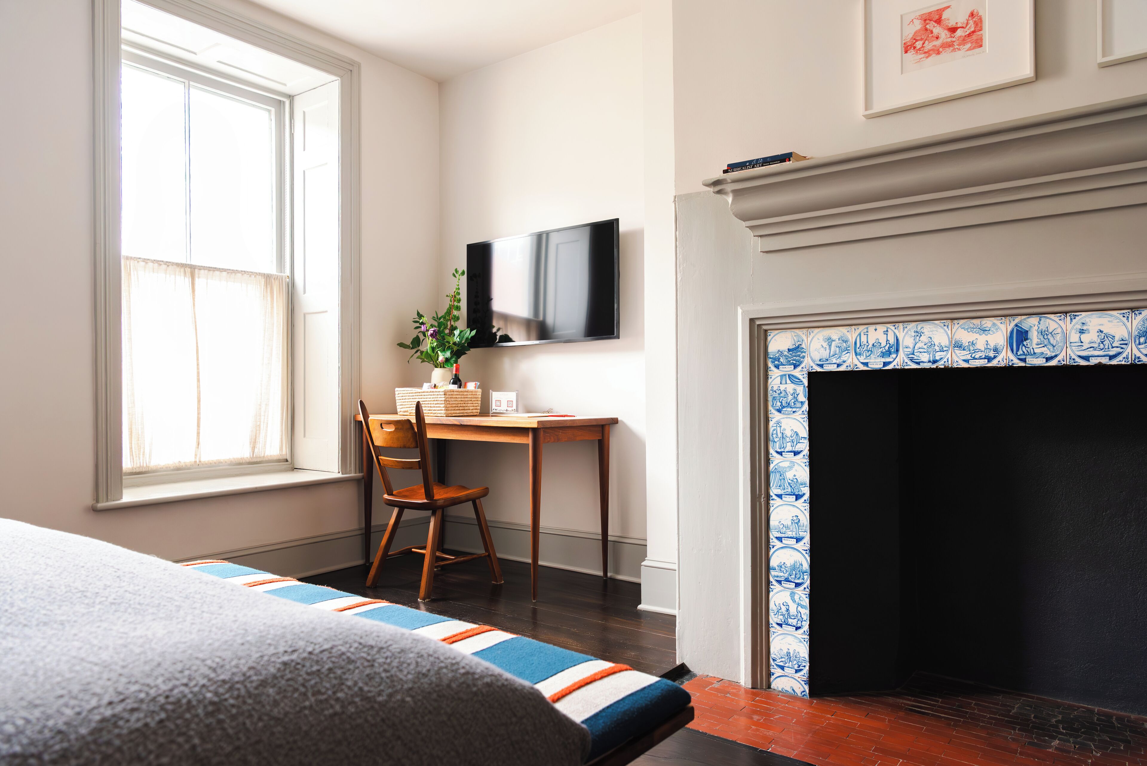 A bedroom with a bed, a desk with a chair, a television, and a fireplace with blue and white tiles.