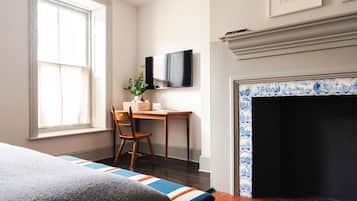 A bedroom with a bed, a desk with a chair, a television, and a fireplace with blue and white tiles.