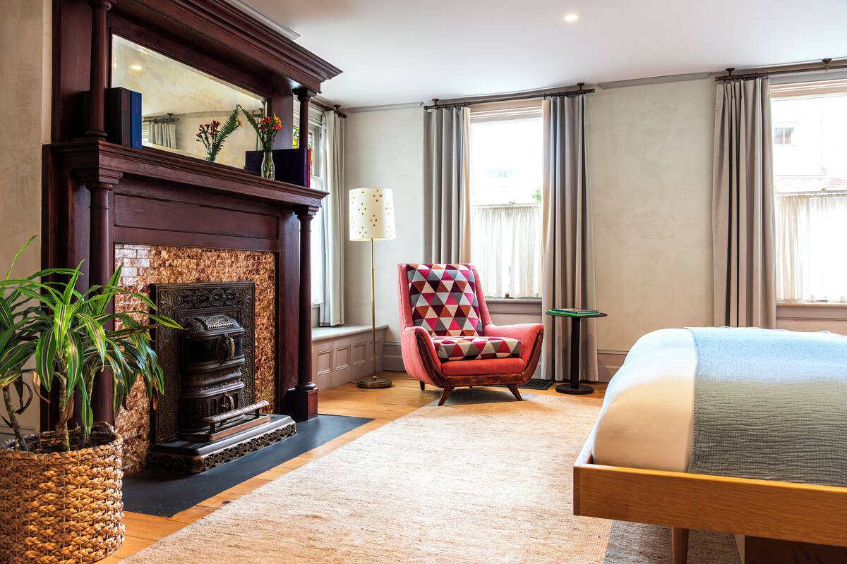 A hotel room with a fireplace, a red armchair, a bed, and large windows.