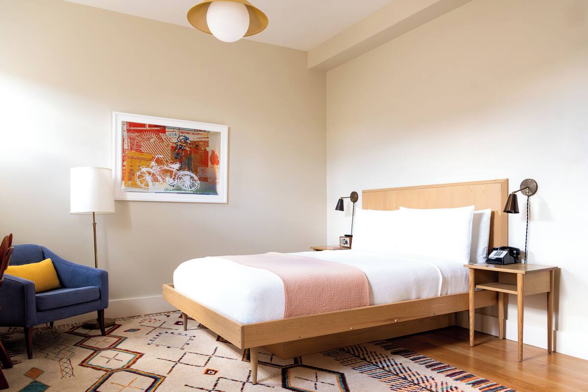 A neatly arranged bedroom with a wooden bed, a blue armchair, a floor lamp, and a wall-mounted artwork.
