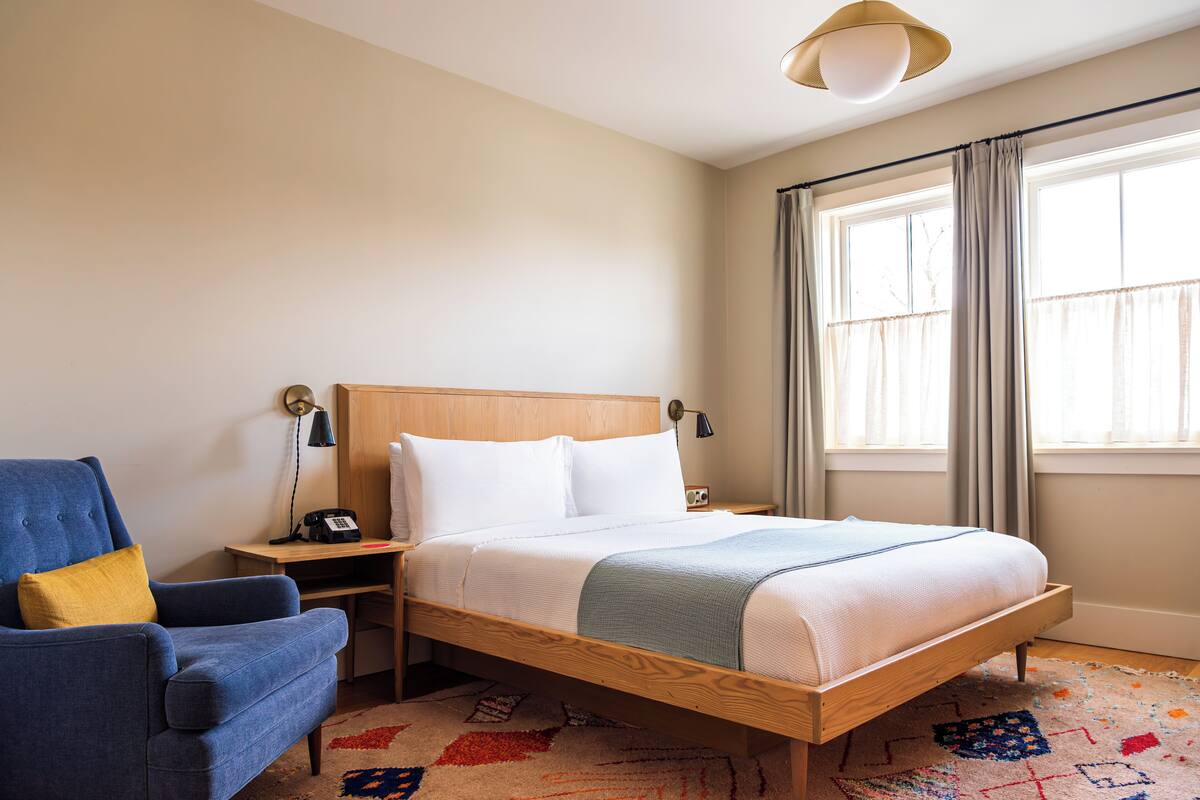 A hotel room with a wooden bed, a blue armchair, a nightstand with a lamp, and a window with curtains.