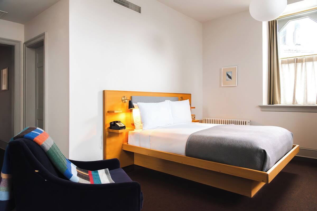 A hotel room with a bed, a chair, a bedside table with a lamp, and a window with curtains.