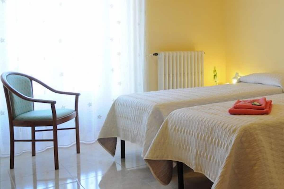 A hotel room with two beds, a chair, a desk with a computer, and a radiator.