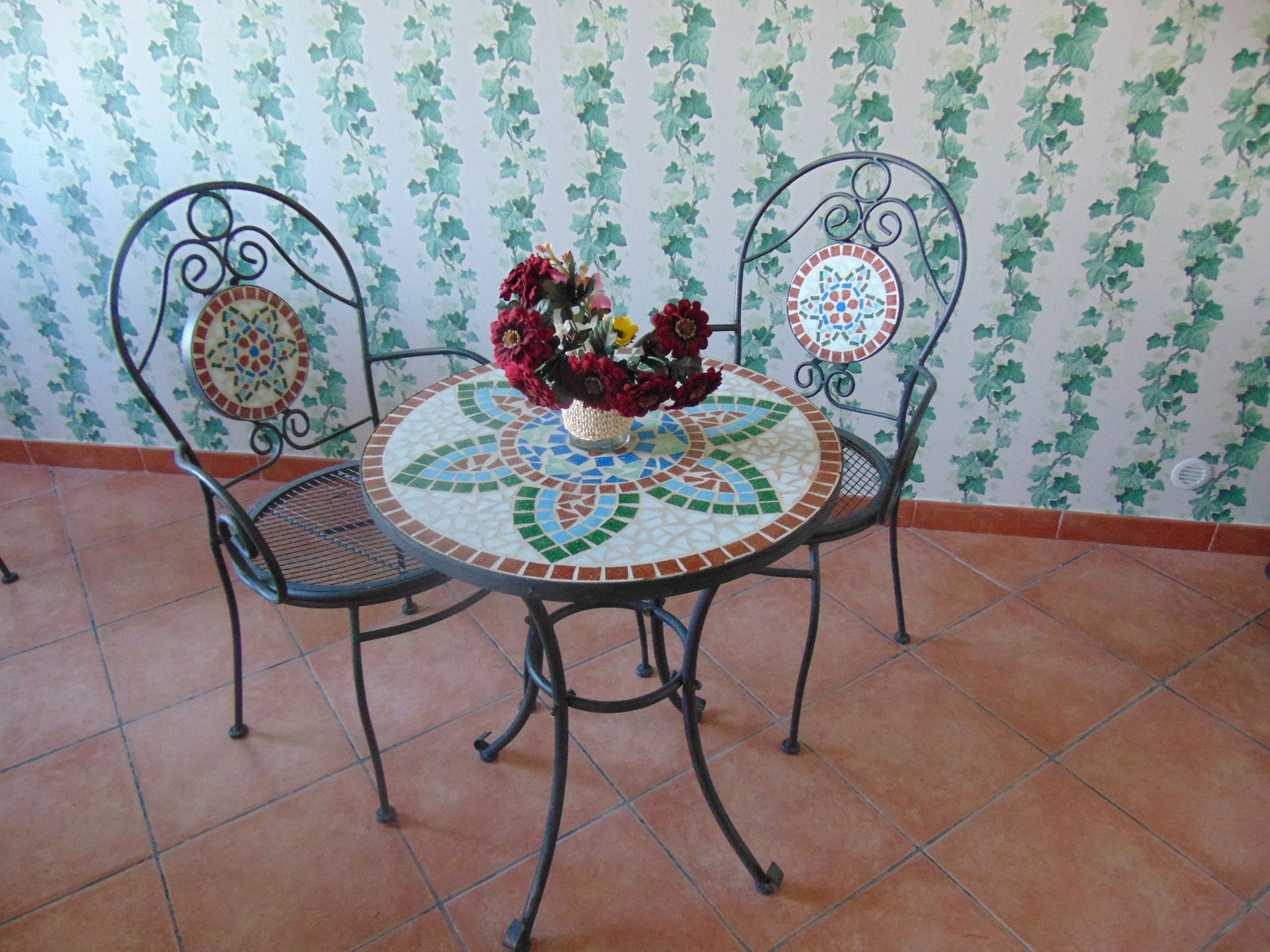 A round table with a mosaic top, two chairs with decorative backs, and a vase with flowers.