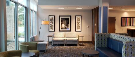 A hotel lobby with a patterned carpet, seating area with sofas and chairs, artwork on the walls, and a large window providing a view outside.