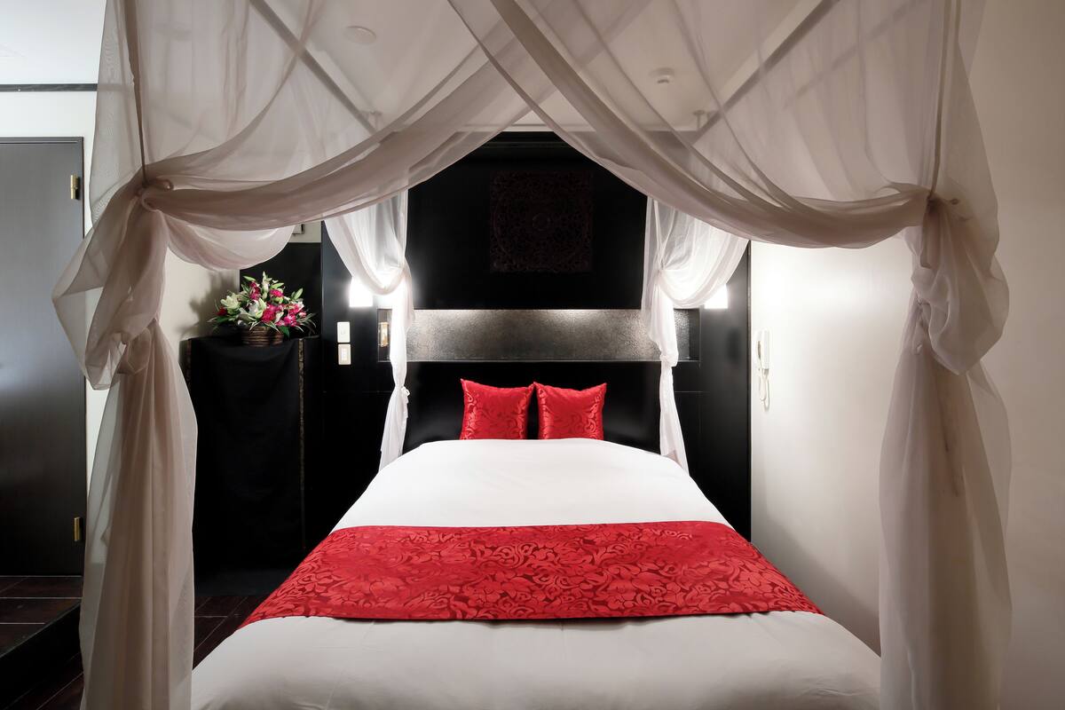 Un lit avec une couvre-lit blanche et rouge, un baldaquin et une table de chevet ornée d’un vase de fleurs.