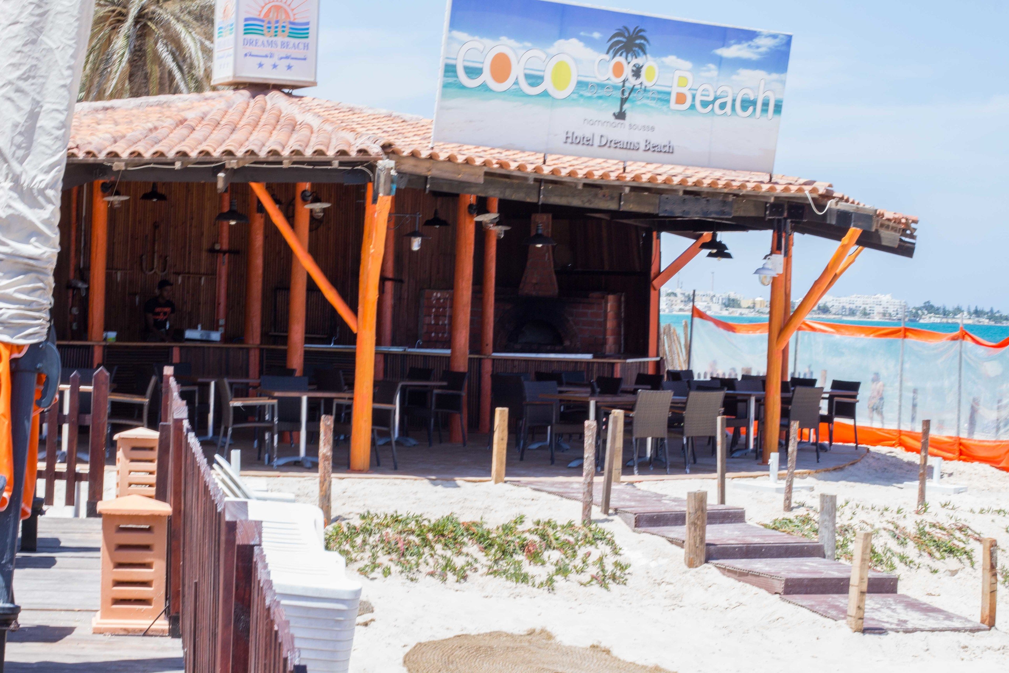 A beachside restaurant with outdoor seating, a sign for Hotel Dreams Beach, and a view of the ocean.