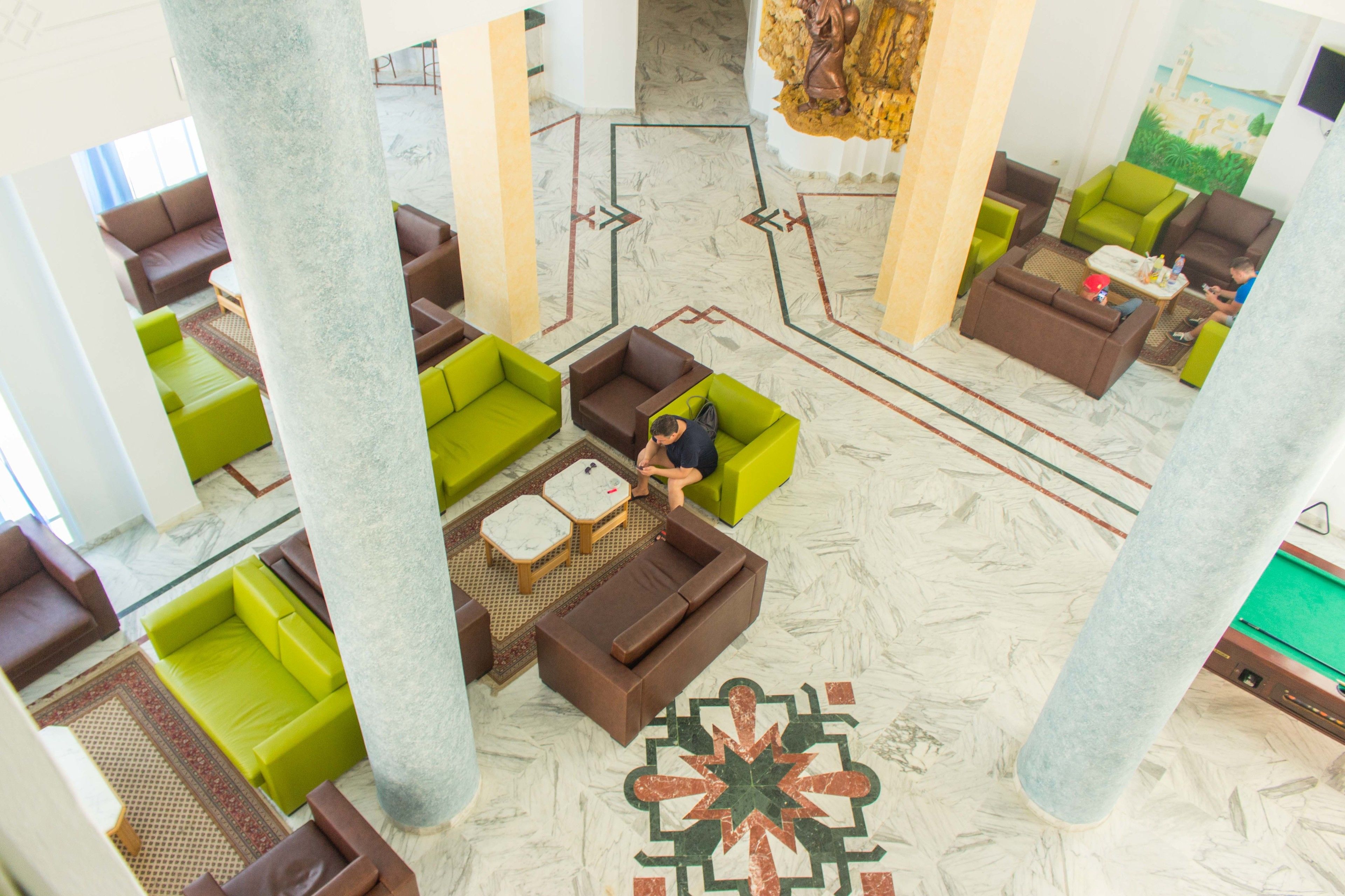 A spacious lobby with multiple seating areas, including green and brown sofas, a coffee table, and a patterned floor.