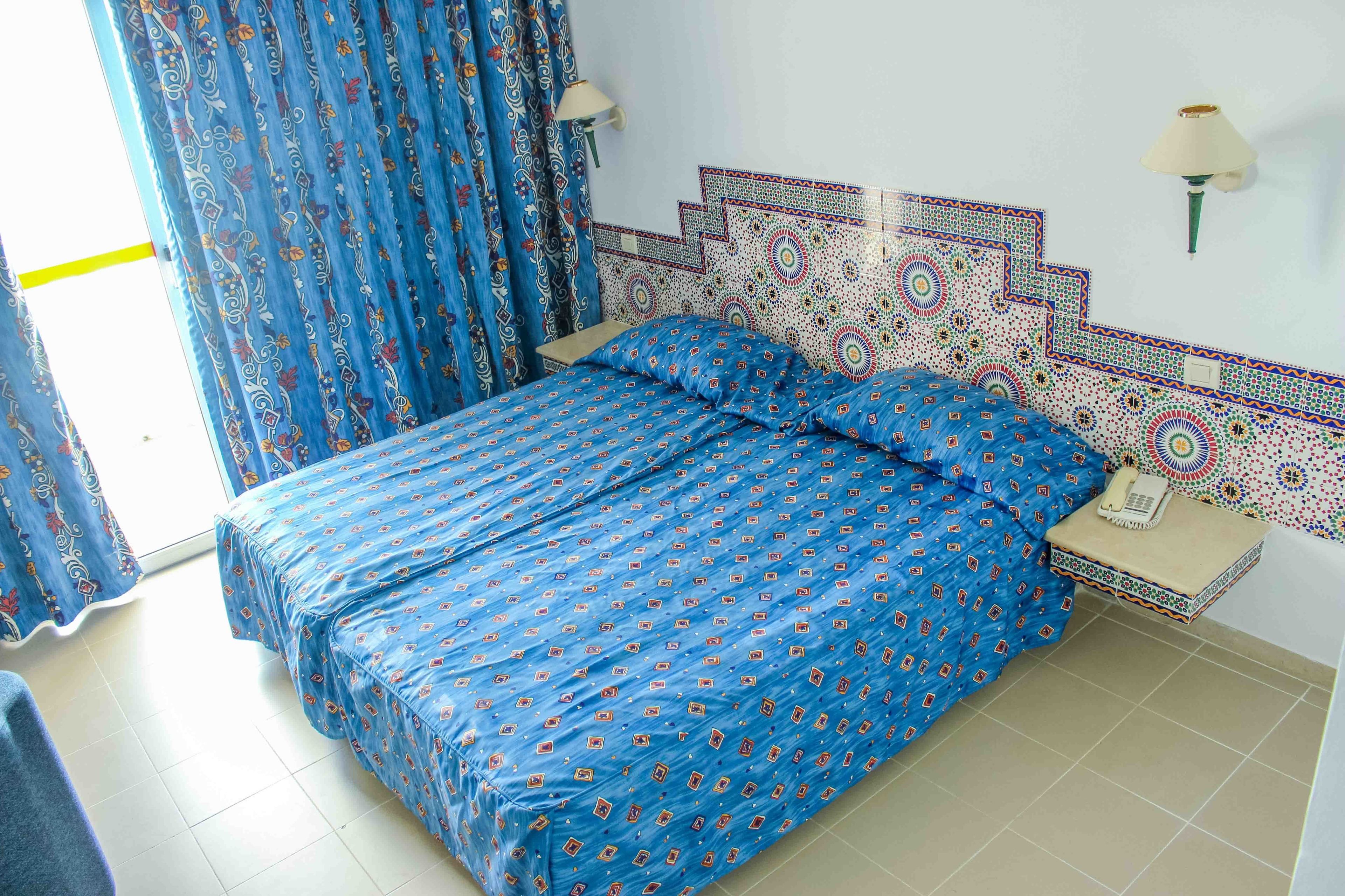 A room with a bed, a nightstand, a lamp, and patterned curtains.