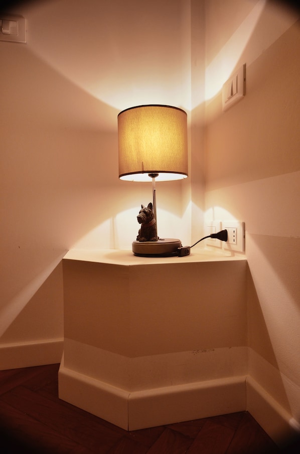 Mesa no canto com luminária e uma pequena estatueta, tomada e parede branca.