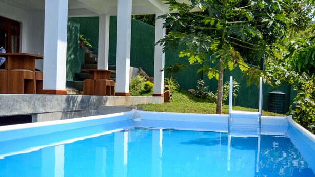 Una piscina con agua cristalina azul, rodeada por un porche cubierto con muebles de madera y una pared verde.