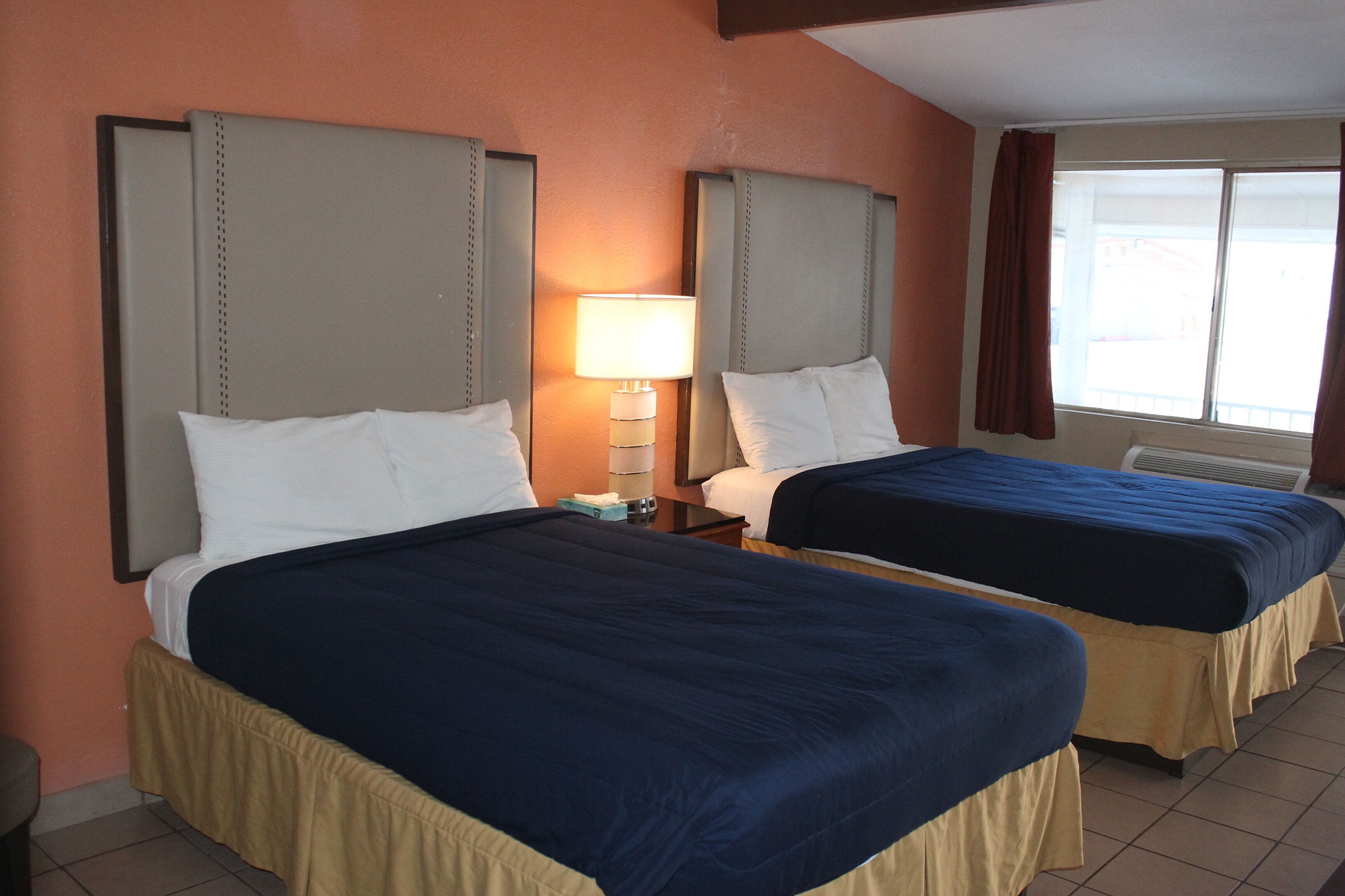 Habitación de hotel con dos camas, cortinas color beige, una ventana con vistas y una lámpara de noche.
