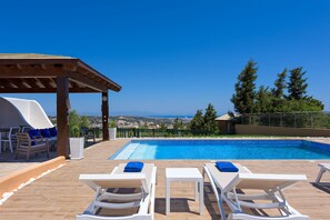 A poolside area with a view of the sea, wooden deck, lounge chairs, and a covered seating area.
