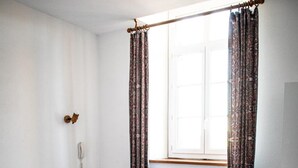 Una habitación pequeña y limpia con dos camas individuales, una mesa de madera, una silla, un radiador y una ventana con cortinas.
