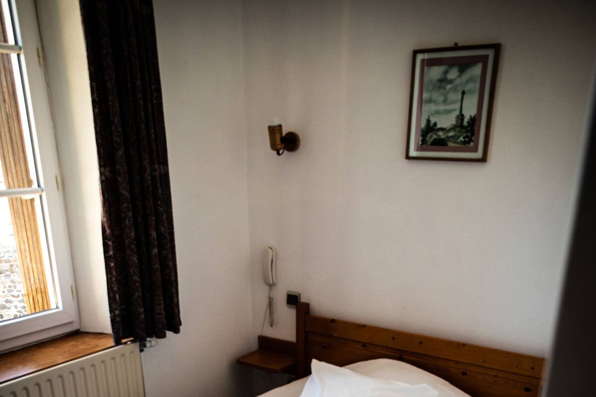 A neatly made bed with white bedding in a room with a wooden headboard, a window with curtains, and a wall-mounted telephone.
