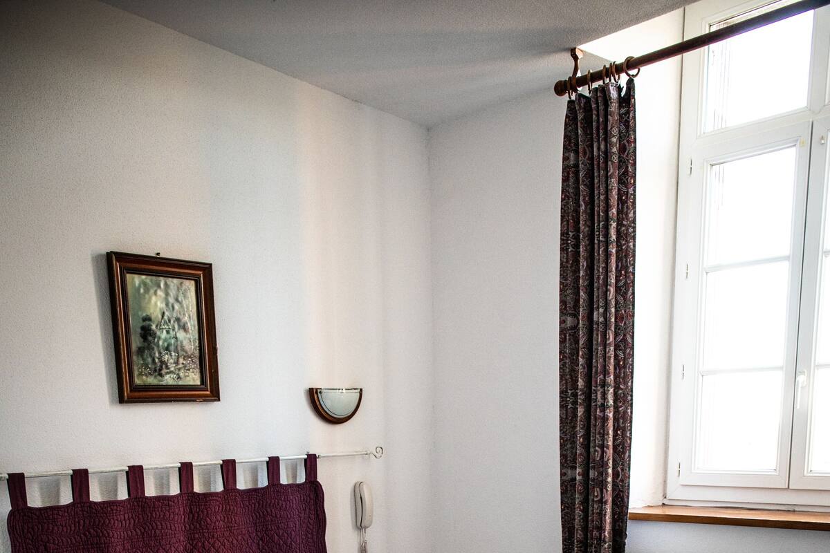 A bedroom with a single bed, a bedside table, a nightstand, a wall-mounted telephone, and a window with curtains.