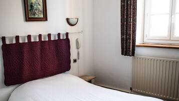 A bedroom with a single bed, a bedside table, a nightstand, a wall-mounted telephone, and a window with curtains.